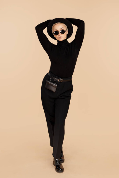 Trendy hipster woman with blonde short hair wearing black stylish clothes, hat and sunglasses over beige background. - 写真・画像