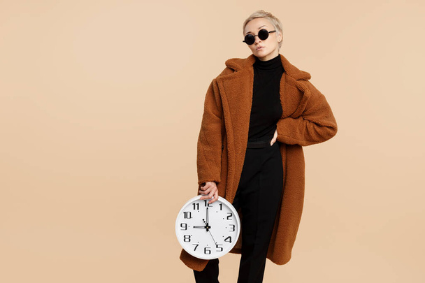 Serious young hipster woman with blonde short hair wearing a coat and sunglasses holding a clock over beige background. - Fotografie, Obrázek