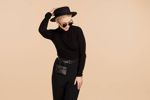 Charming hipster woman with blonde short hair wearing black stylish clothes, hat and sunglasses over beige background. - Fotó, kép