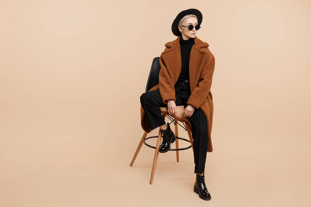 Serious young hipster woman with blonde short hair wearing a coat, hat and sunglasses posing over beige background. - 写真・画像