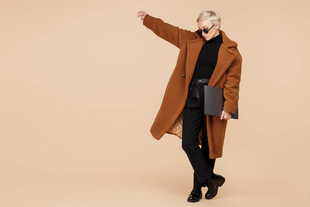 Trendy young hipster woman with blonde short hair wearing a coat and sunglasses holding computer over beige background. - Foto, Bild