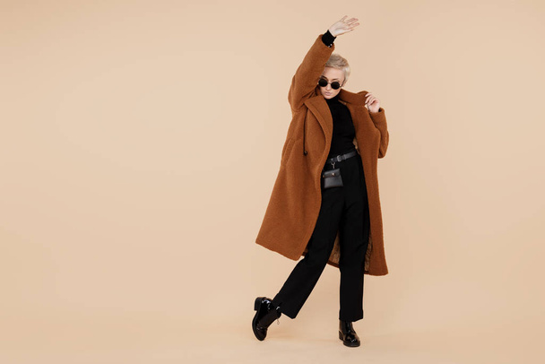 Happy young hipster woman with blonde short hair wearing a coat and sunglasses dance over beige background. Copy space. - Valokuva, kuva