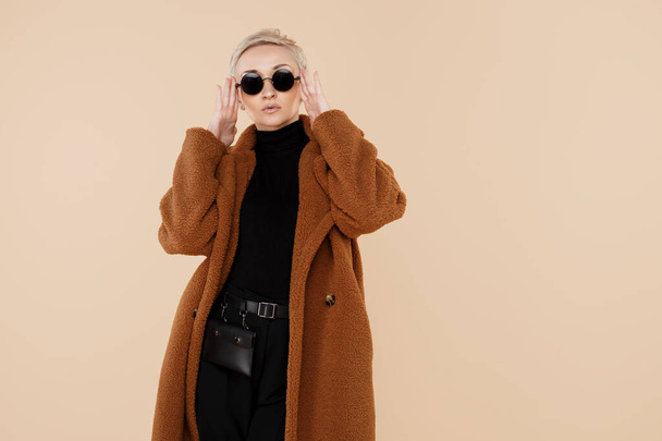 Happy young hipster woman with blonde short hair wearing a coat and sunglasses posing over beige background. - Photo, Image