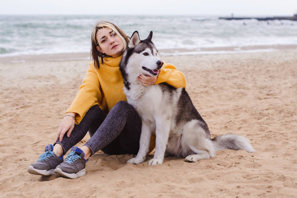 Молода красива жінка в жовтому светрі грає з собакою (сірий і білий хаскі) на пляжі
 - Фото, зображення