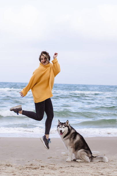 Młoda piękna kobieta w żółtym swetrze z pulowerem bawi się na plaży z psem (szaro-białym husky) - Zdjęcie, obraz