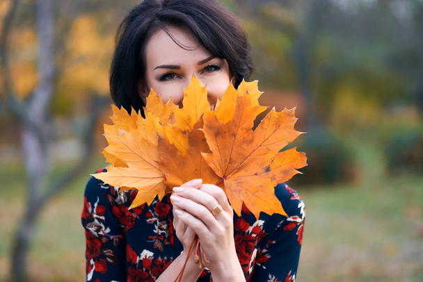 Beautiful elegant woman standing and posing with bouquet of yellow leaves in autumn city park, face closeup - Foto, afbeelding