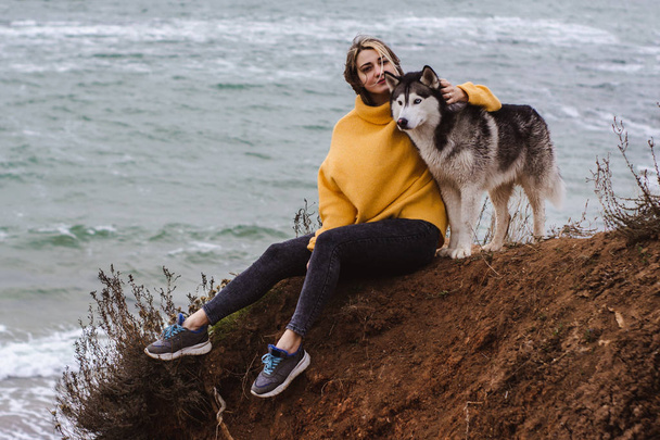 junge schöne Frau im gelben Pullover spielt mit einem Hund (graue und weiße Huskys) am Strand - Foto, Bild