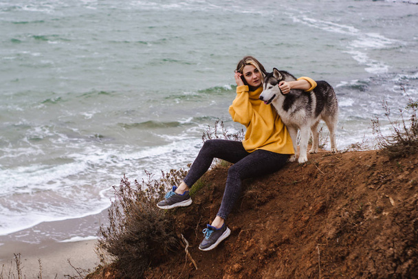 Young beautiful woman in yellow pullover sweater plays with a dog (grey and white husky) on the beach - 写真・画像