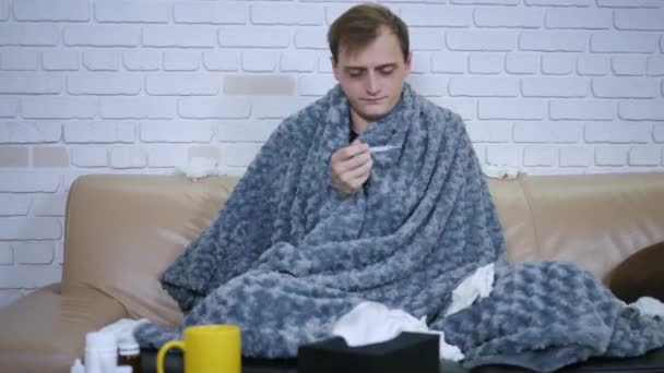 Joven hombre caucásico se siente enfermo sentado en el sofá y luego comprobar la temperatura corporal utilizando un termómetro. Enfermedad persona enfermedad virus retrato
 - Imágenes, Vídeo