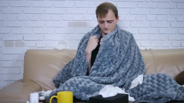 Молодой кавказский мужчина плохо себя чувствует, сидя на диване, а затем проверяет температуру тела с помощью термометра. Портрет вируса болезни человека
. - Кадры, видео