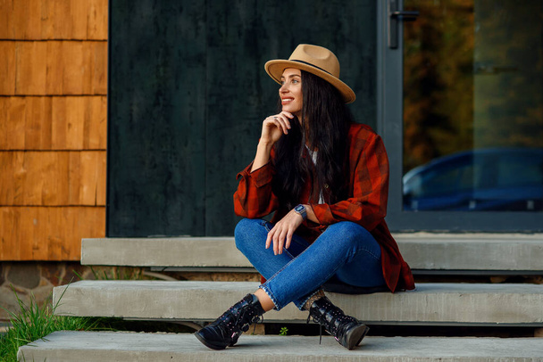 Beautiful woman in a red shirt, jeans and hat sits on the stairs of her modern home near the forest. - Foto, afbeelding