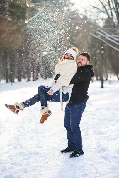 Young stylish couple in love have fun while walking on the snowy winter park. Handsome young man holds his beautiful girl on the hands. - Foto, imagen
