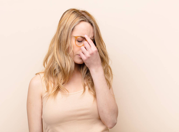 young pretty blonde woman feeling stressed, unhappy and frustrated, touching forehead and suffering migraine of severe headache against flat color wall - Photo, Image