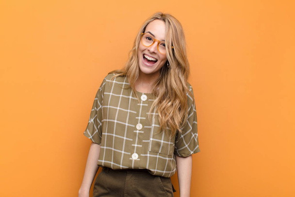 young pretty blonde woman with a big, friendly, carefree smile, looking positive, relaxed and happy, chilling against flat color wall - Photo, Image