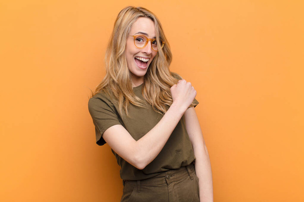 young pretty blonde woman feeling happy, positive and successful, motivated when facing a challenge or celebrating good results against flat color wall - Foto, Imagem
