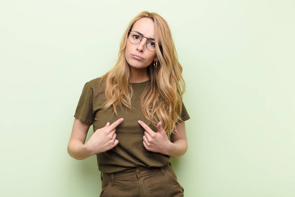 young pretty blonde woman pointing to self with a confused and quizzical look, shocked and surprised to be chosen against flat color wall - Foto, Imagen