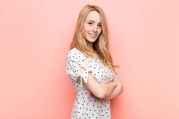 young pretty blonde woman smiling to camera with crossed arms and a happy, confident, satisfied expression, lateral view against flat color wall - Foto, Bild