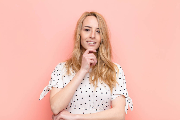 young pretty blonde woman looking happy and smiling with hand on chin, wondering or asking a question, comparing options against flat color wall - Фото, зображення