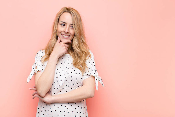 young pretty blonde woman smiling with a happy, confident expression with hand on chin, wondering and looking to the side against flat color wall - Photo, Image