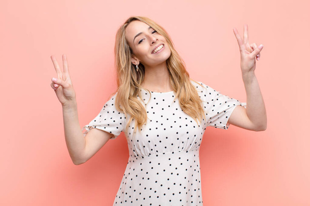 young pretty blonde woman smiling and looking happy, friendly and satisfied, gesturing victory or peace with both hands against flat color wall - Fotografie, Obrázek
