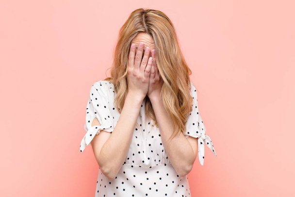 young pretty blonde woman feeling sad, frustrated, nervous and depressed, covering face with both hands, crying against flat color wall - Foto, Bild