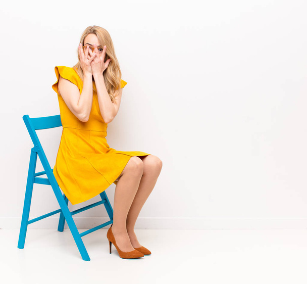 young pretty blonde woman feeling scared or embarrassed, peeking or spying with eyes half-covered with hands against flat color wall - Foto, Imagen