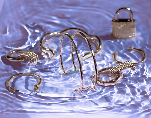 Χρυσά βραχιόλια, σκουλαρίκια, δαχτυλίδια, κοσμήματα σε μωβ νερό πίσω - Φωτογραφία, εικόνα