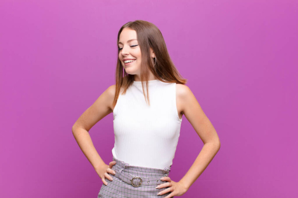 young red head woman looking happy, cheerful and confident, smiling proudly and looking to side with both hands on hips against flat wall - Photo, image