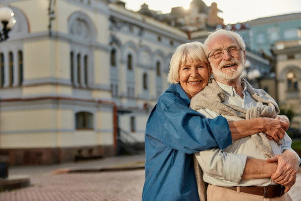 Pravá láska nemá žádné datum spotřeby. Portrét veselého staršího páru v neformálním oblečení objímající se a dívající se na fotoaparát s úsměvem, zatímco spolu stojí venku - Fotografie, Obrázek