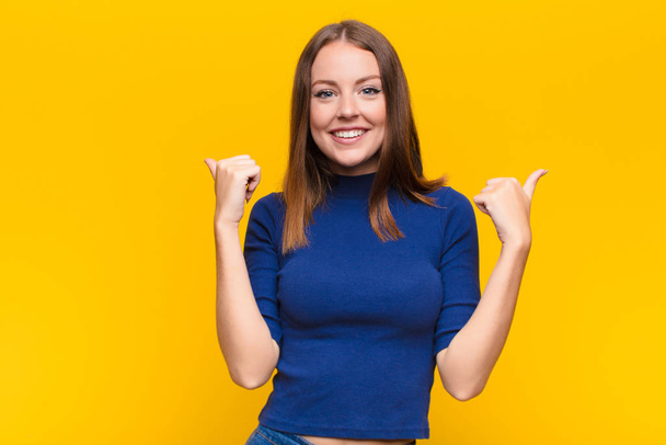 young red head woman smiling joyfully and looking happy, feeling carefree and positive with both thumbs up against flat wall - Foto, Bild