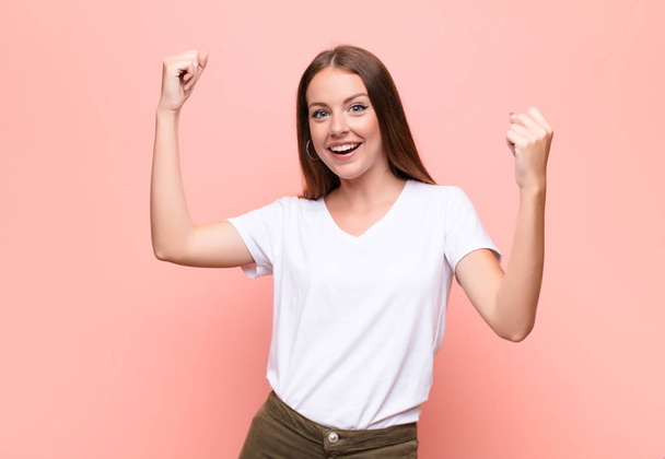 young red head woman shouting triumphantly, looking like excited, happy and surprised winner, celebrating against flat wall - 写真・画像