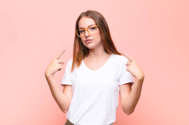 young red head woman with a bad attitude looking proud and aggressive, pointing upwards or making fun sign with hands against flat wall - Foto, imagen