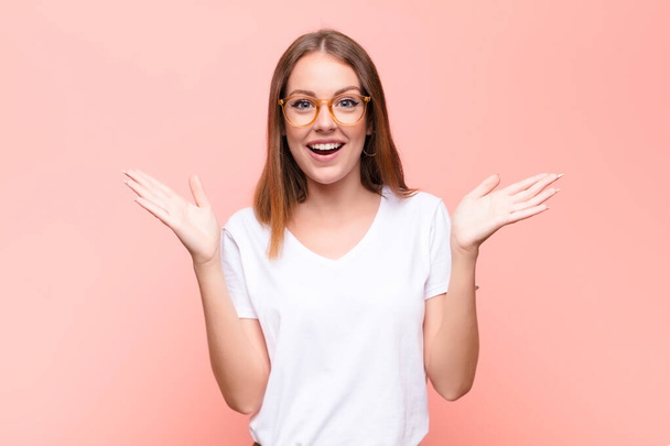 young red head woman looking happy and excited, shocked with an unexpected surprise with both hands open next to face against flat wall - 写真・画像
