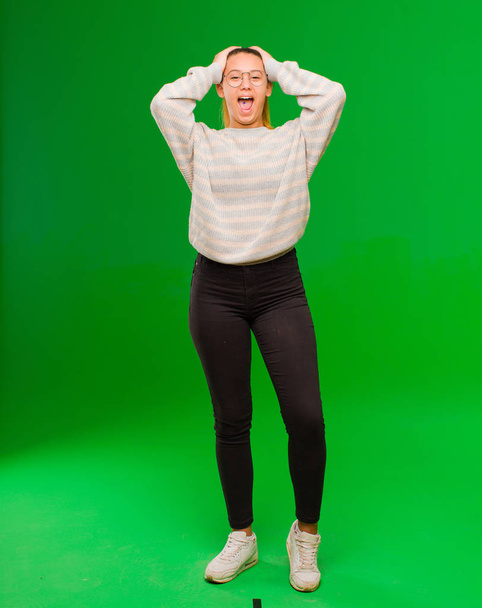 young pretty latin woman raising hands to head, open-mouthed, feeling extremely lucky, surprised, excited and happy against green wall - Foto, imagen