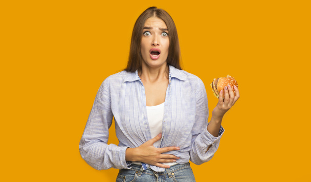 Mädchen hält Burger mit Bauchschmerzen auf gelbem Studiohintergrund - Foto, Bild