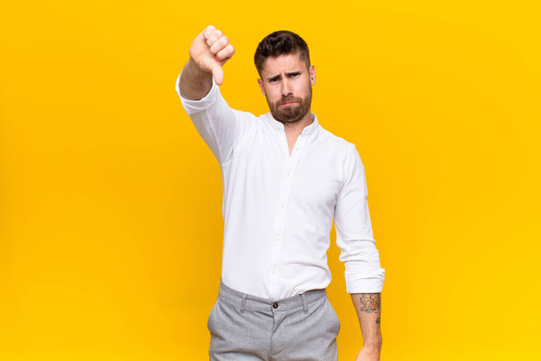 young handosme man feeling cross, angry, annoyed, disappointed or displeased, showing thumbs down with a serious look against flat color wall - Foto, Bild