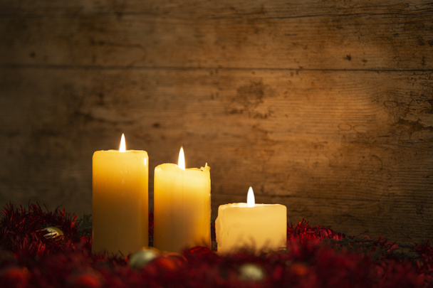 A karácsonyi koncepció melege: három gyertya világít egy könnyű fa asztalon és rusztikus környezetben fenyőágakkal, vörös dekoráció és arany és piros fényes baubles bokeh hatással - Fotó, kép