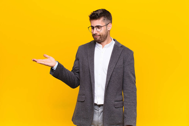 young handosme man feeling happy and smiling casually, looking to an object or concept held on the hand on the side against flat color wall - Foto, imagen