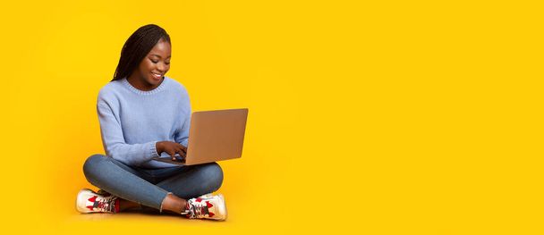 Glimlachend zwart meisje met behulp van laptop over gele achtergrond - Foto, afbeelding