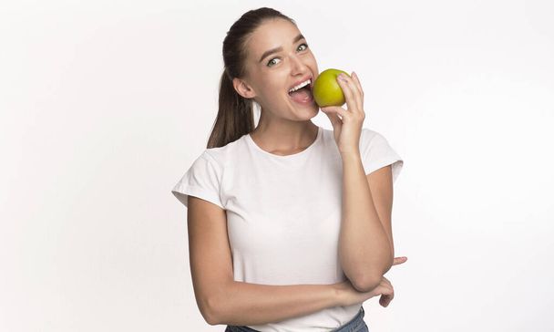 vrouw bijten appel kijken naar camera staande in studio - Foto, afbeelding