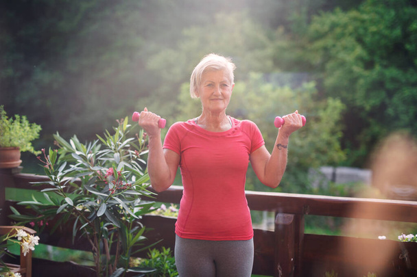 A senior woman standing outdoors on a terrace in summer, doing exercise. - Photo, image