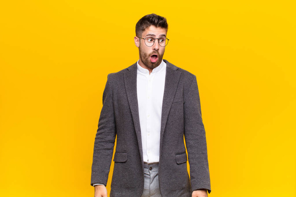 young handosme man feeling shocked, happy, amazed and surprised, looking to the side with open mouth against flat color wall - Фото, изображение