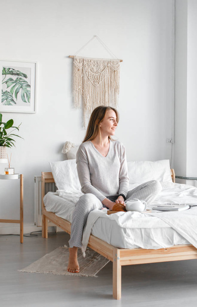 Rest after a hard days work. Beautiful young smiling business woman in pajamas drinking coffee and looking away while sitting on a bed in a white room. - Photo, Image