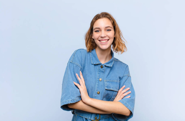 young blonde woman looking like a happy, proud and satisfied achiever smiling with arms crossed against flat color wall - Foto, Bild