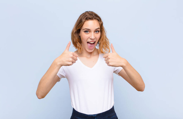 young blonde woman smiling broadly looking happy, positive, confident and successful, with both thumbs up against flat color wall - Фото, изображение
