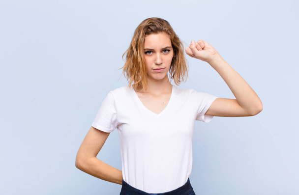 young blonde woman feeling serious, strong and rebellious, raising fist up, protesting or fighting for revolution against flat color wall - Фото, изображение