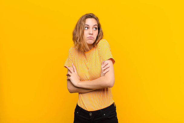 young blonde woman shrugging, feeling confused and uncertain, doubting with arms crossed and puzzled look against flat color wall - Foto, imagen