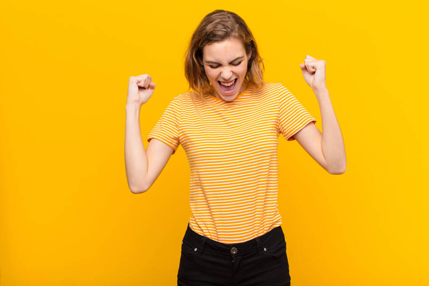 若いブロンドの女性は非常に幸せそして驚き成功を祝いフラットカラーの壁に向かって叫び飛び跳ねる - 写真・画像