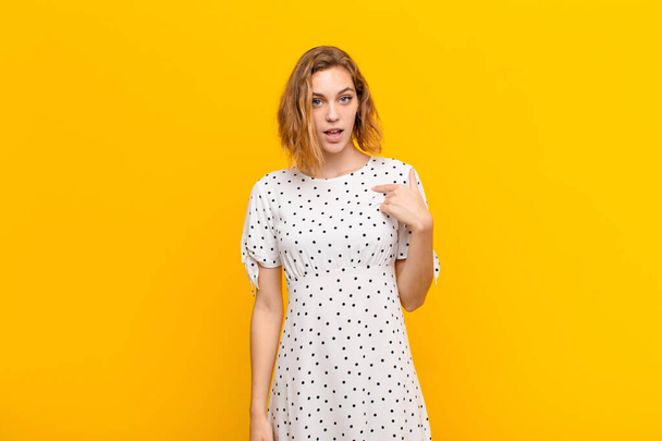 young blonde woman looking shocked and surprised with mouth wide open, pointing to self against flat color wall - Φωτογραφία, εικόνα