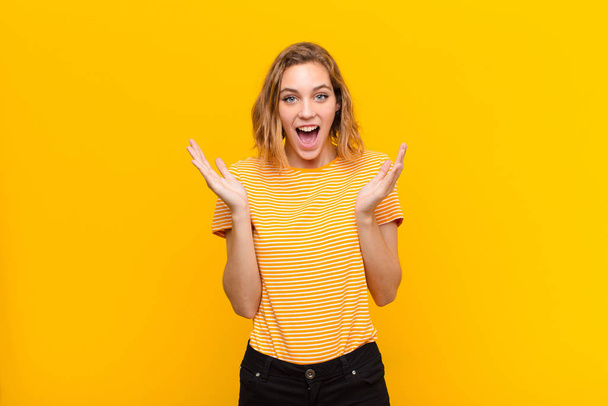young blonde woman feeling happy, excited, surprised or shocked, smiling and astonished at something unbelievable against flat color wall - Photo, Image
